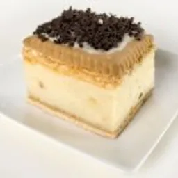 Kwadratowy kawałek domowego ciasta kremowego na białym, kwadratowym talerzu, udekorowany drobnymi wiórkami czekoladowymi.