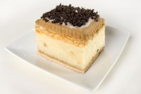 Kwadratowy kawałek domowego ciasta kremowego na białym, kwadratowym talerzu, udekorowany drobnymi wiórkami czekoladowymi.