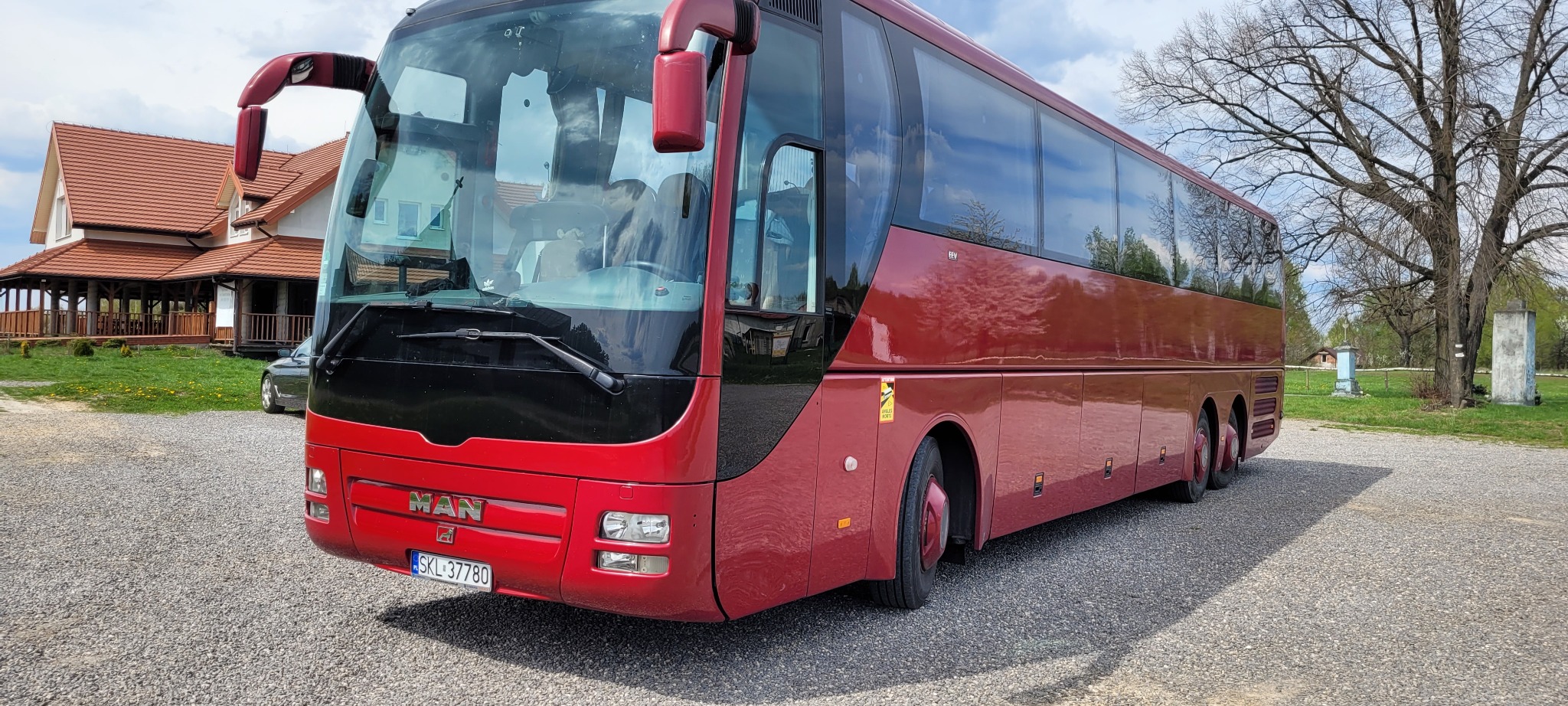 Czerwony autokar MAN na żwirowym parkingu przed budynkiem z czerwoną dachówką.