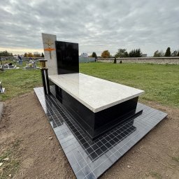 Zakład Kamieniarski Ireneusz Baran - Nowoczesny nagrobek z jasnego i czarnego kamienia na cmentarzu, z krzyżem i lampionem. Tło stanowią inne nagrobki i mur z drzewami.
