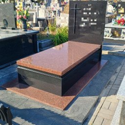 Zakład Kamieniarski Ireneusz Baran - Granitowy nagrobek w odcieniach czerni i czerwieni na cmentarzu, z krzyżem i inskrypcją. W tle inne groby i zieleń. Słoneczny dzień.