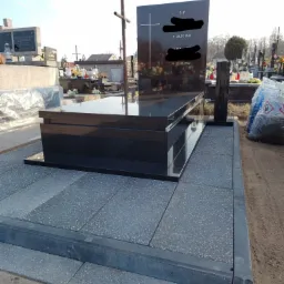 Czarny, błyszczący pomnik z krzyżem na cmentarzu, otoczony szarą kostką brukową i innymi nagrobkami w tle.