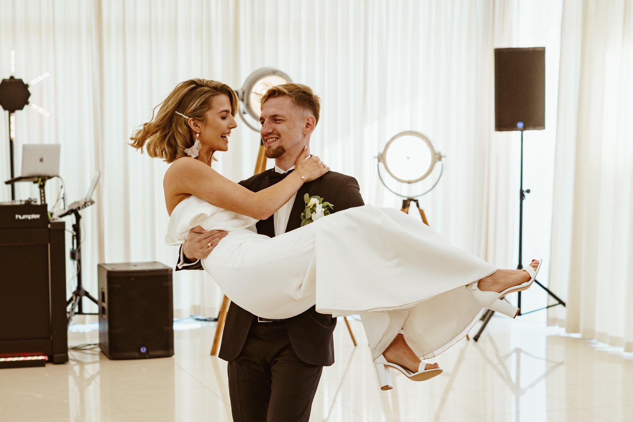Uśmiechnięty pan młody trzyma na rękach uśmiechniętą pannę młodą w białej sukni i białych butach, w tle białe zasłony i oświetlenie sceniczne.