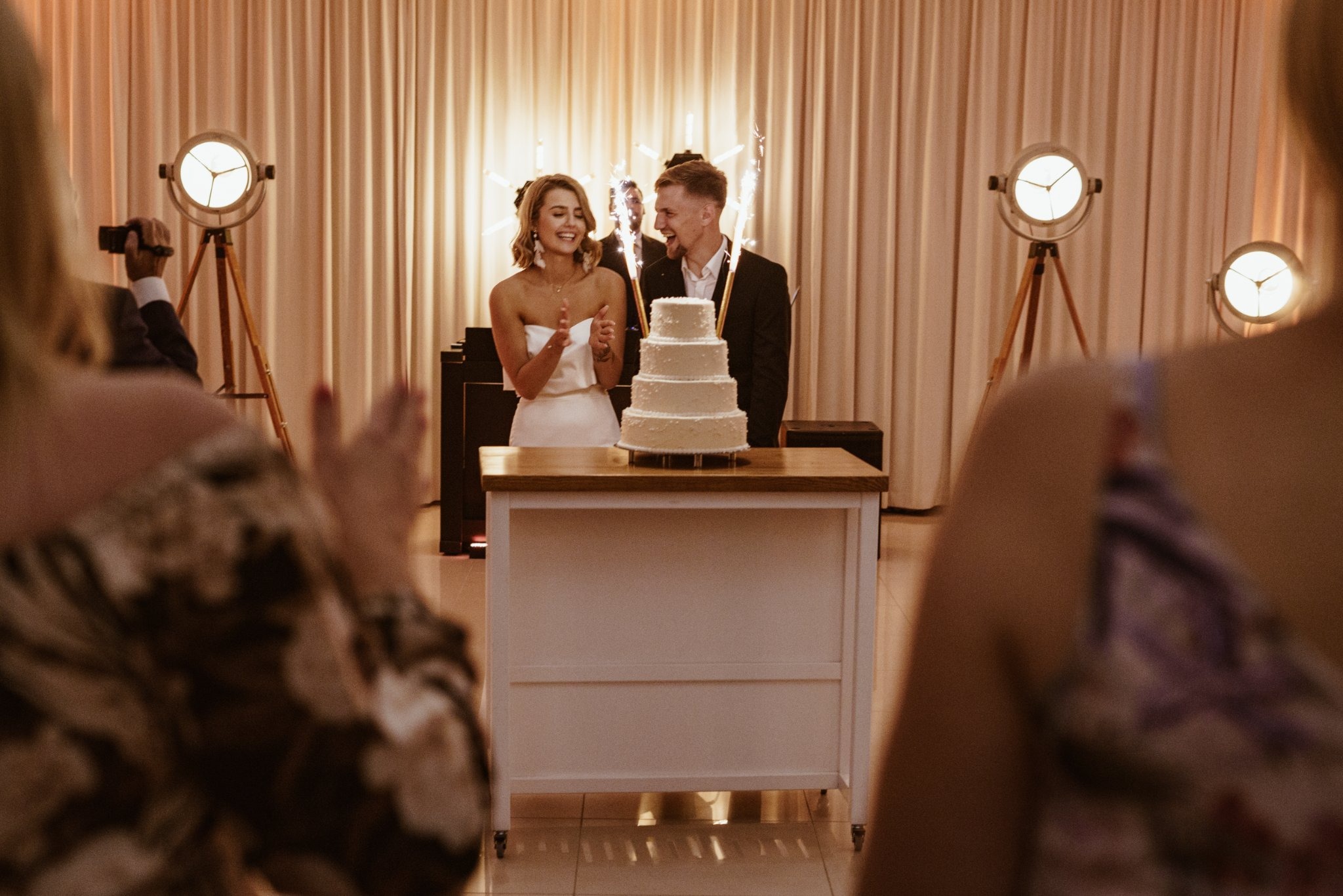 Uśmiechnięta para młoda przy piętrowym torcie weselnym z fontannami iskier, na tle beżowych zasłon i reflektorów studyjnych, z widocznymi osobami w tle.