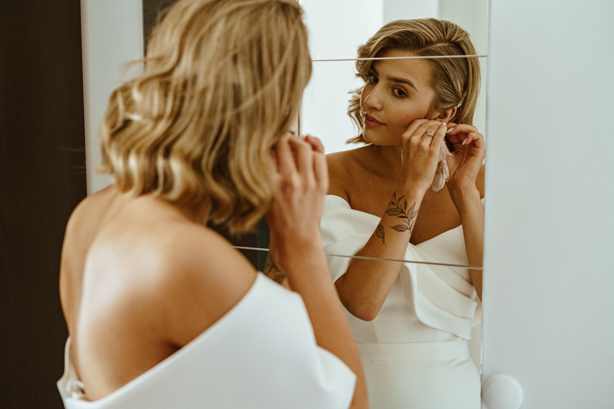 Panna młoda w białej sukni, przeglądająca się w lustrze i zakładająca kolczyk, widoczny tatuaż na ramieniu.