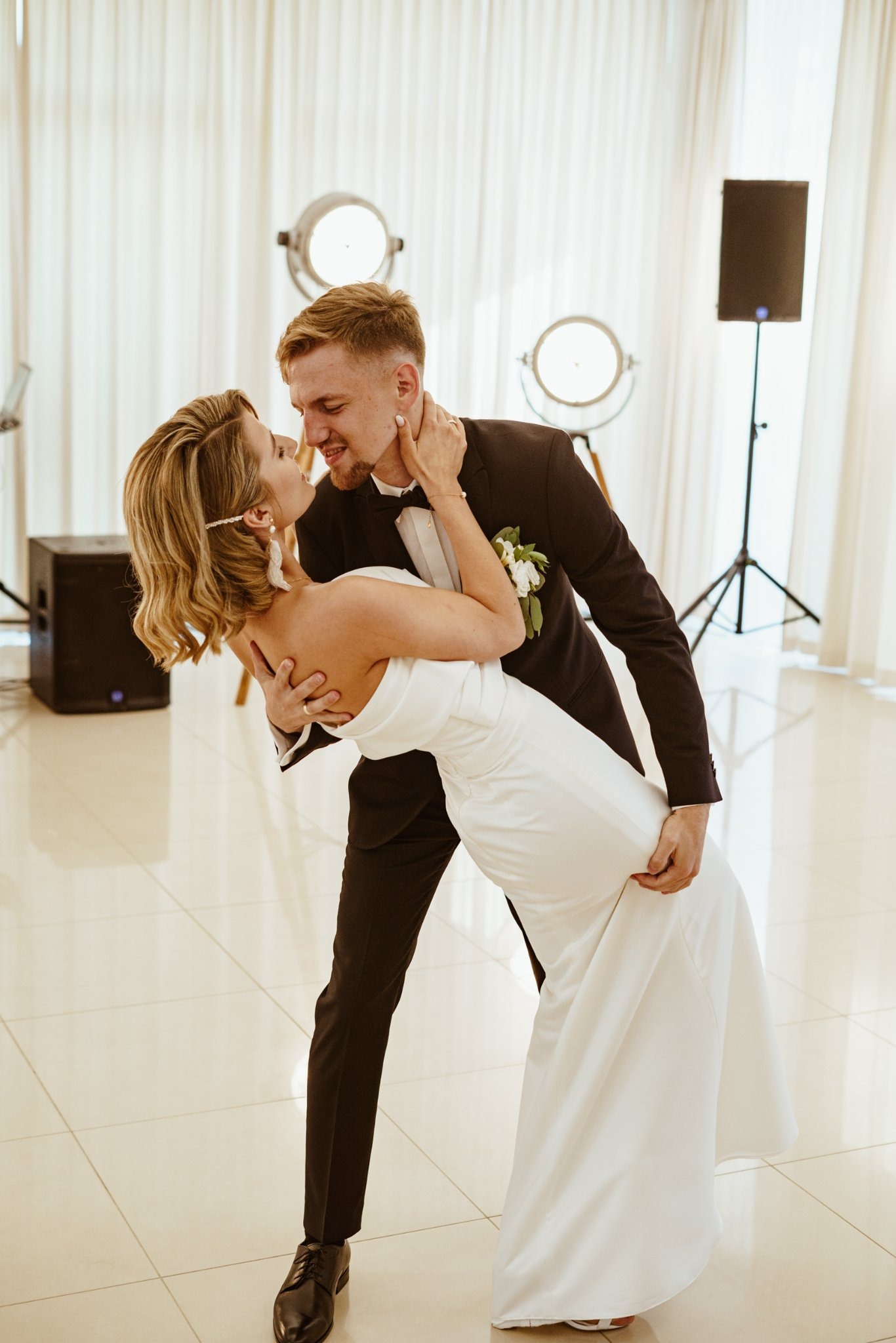 Młoda para w tańcu podczas wesela, panna młoda w białej sukni, pan młody w czarnym smokingu z białą muszką, oświetlenie sceniczne w tle.