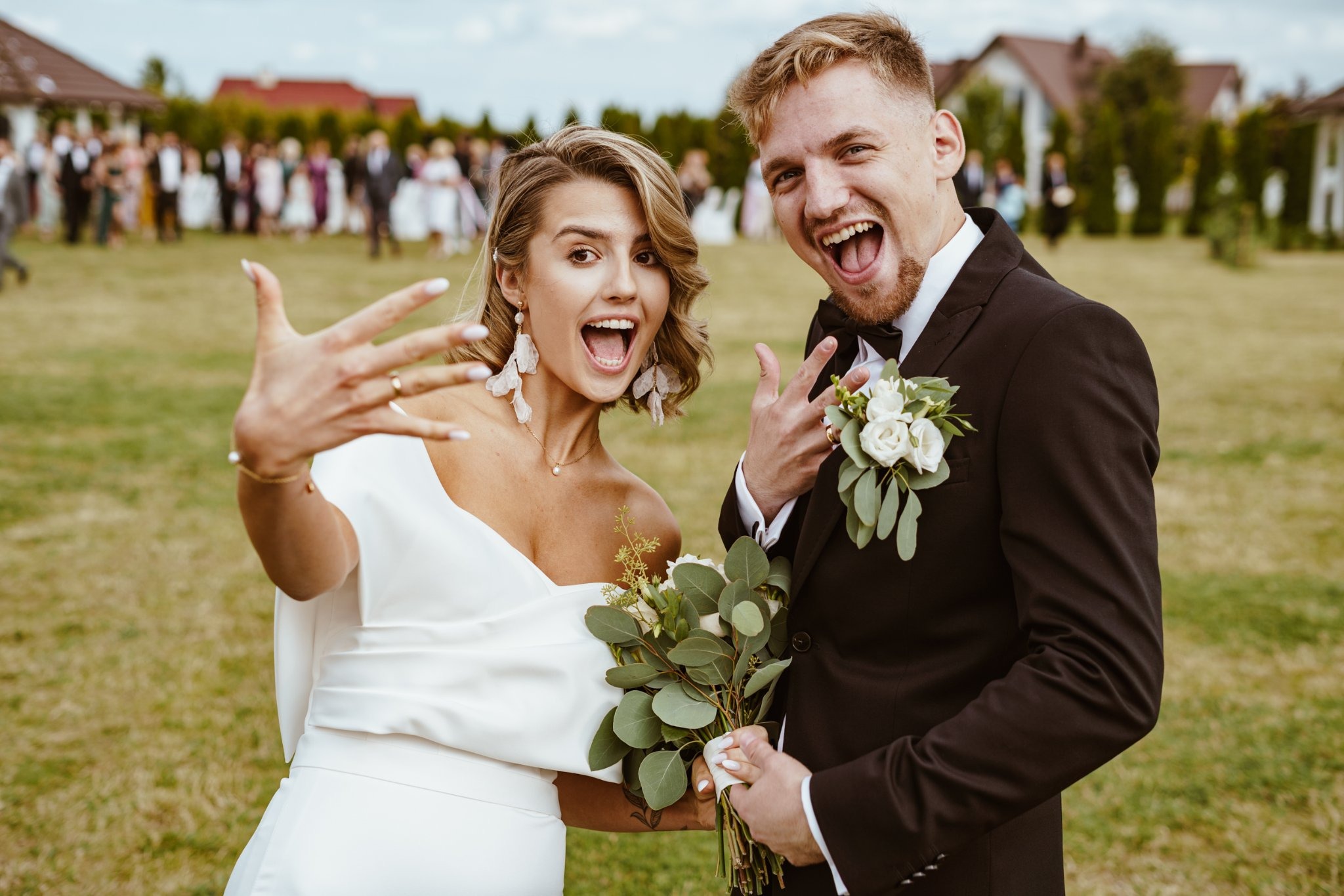 Uśmiechnięta para młoda w plenerze, panna młoda w białej sukni z bukietem i ozdobnymi kolczykami, pan młody w czarnym garniturze z butonierką, oboje radośnie pozują, pokazując obrączki na palcach...