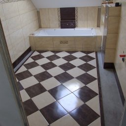 PHU Kambud - Wykończona łazienka na poddaszu z białą wanną i szachownicą z brązowo-beżowych płytek podłogowych, beżowe płytki ścienne z dekoracyjnym wnęką nad wanną.