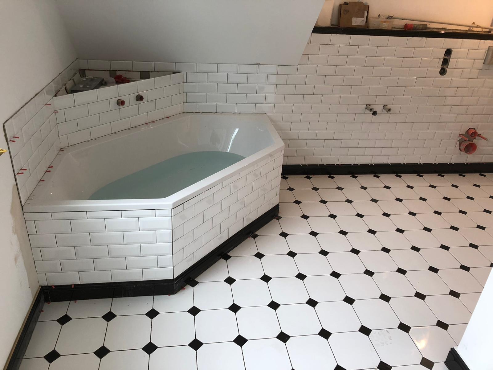 Narożna wanna wypełniona wodą, obłożona białymi, prostokątnymi płytkami z czarną listwą, dopasowana do podłogi w biało-czarną szachownicę; widoczne elementy instalacji na ścianie i dystanse do płytek.