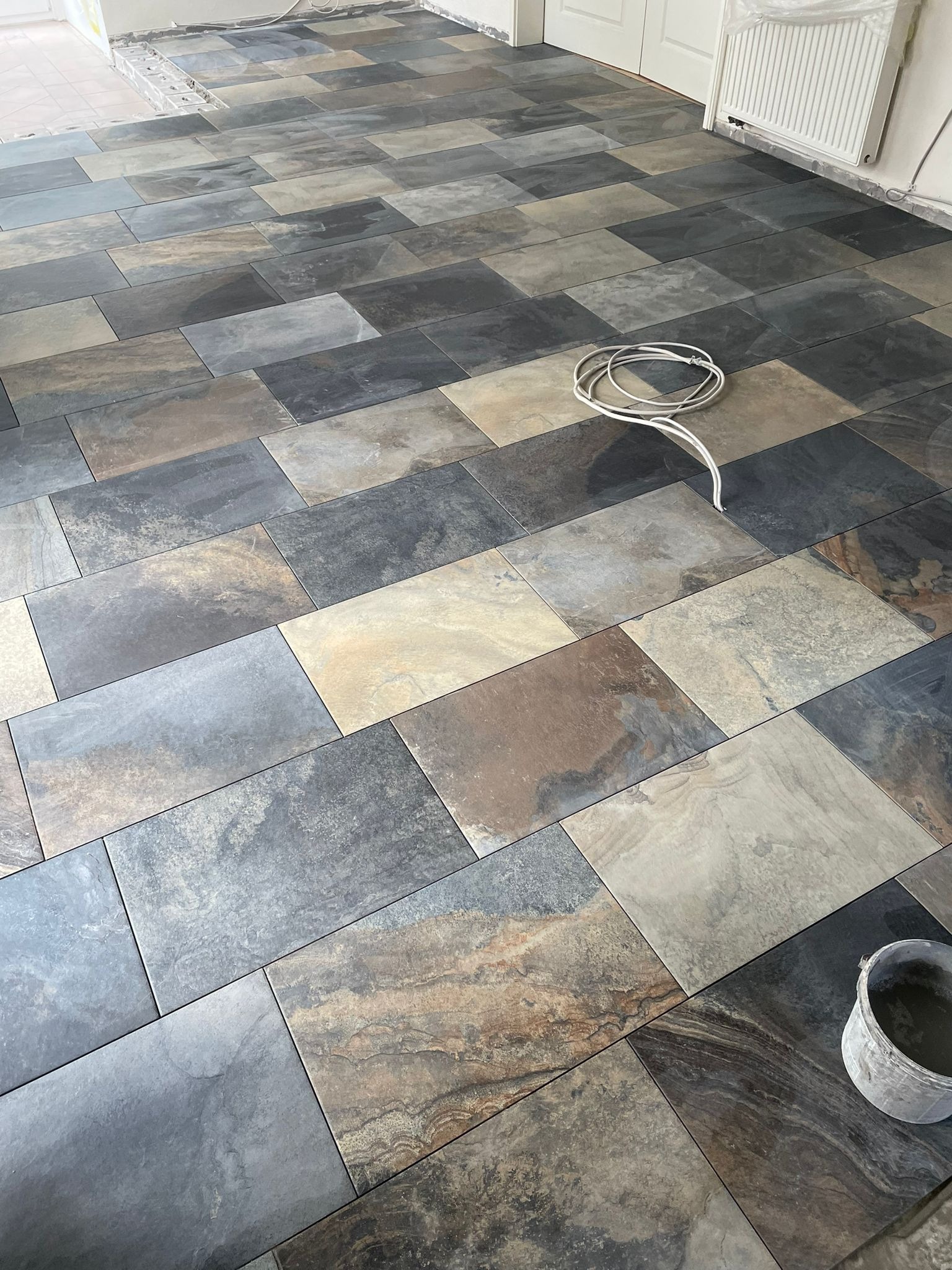 Ułożona podłoga z płytek imitujących kamień w odcieniach szarości, beżu i brązu; widoczny kabel elektryczny i wiadro z zaprawą.