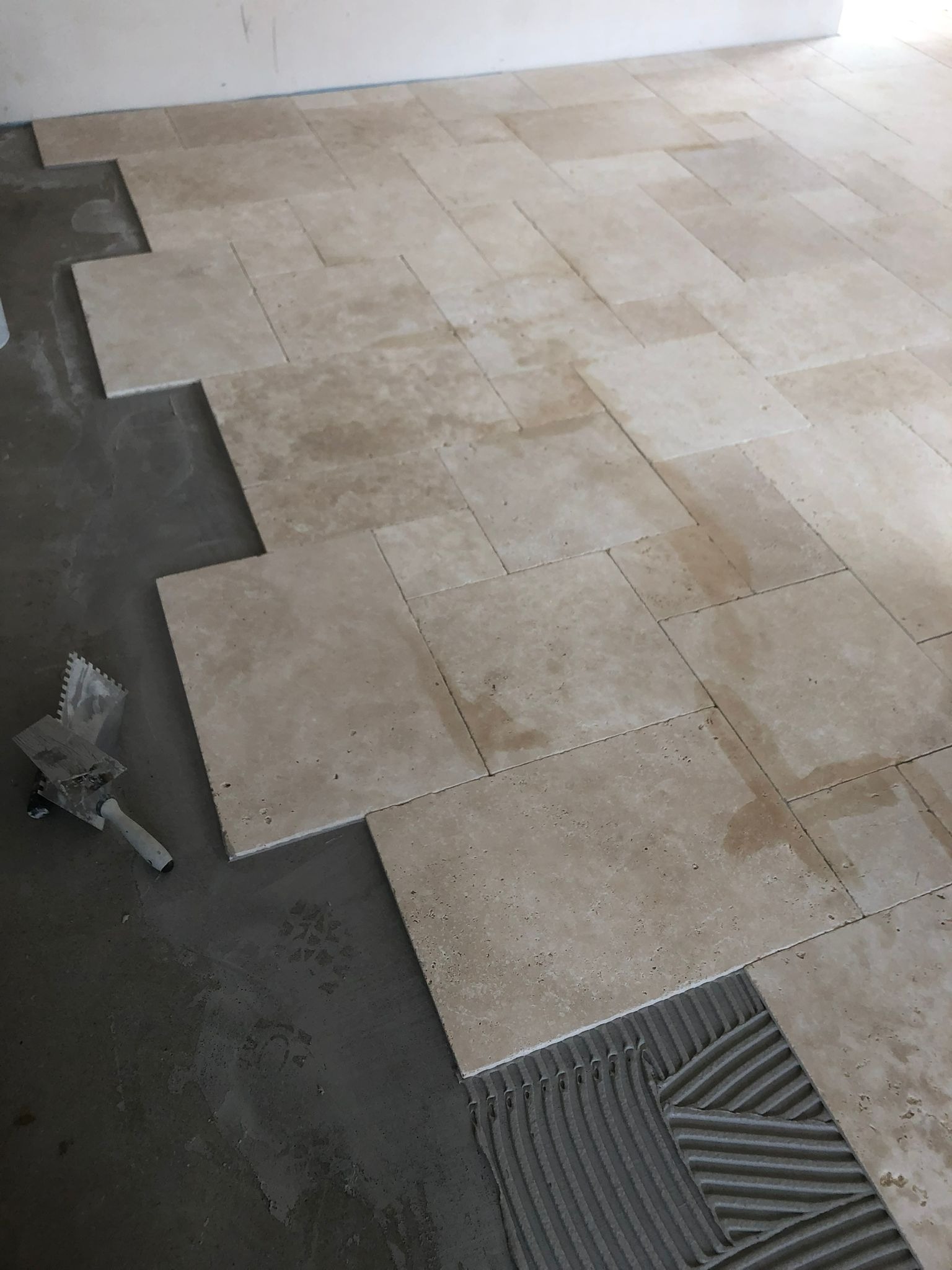 Układanie beżowych płytek podłogowych na zaprawie, widoczna paca z resztkami kleju, fragment nieukończonej podłogi.