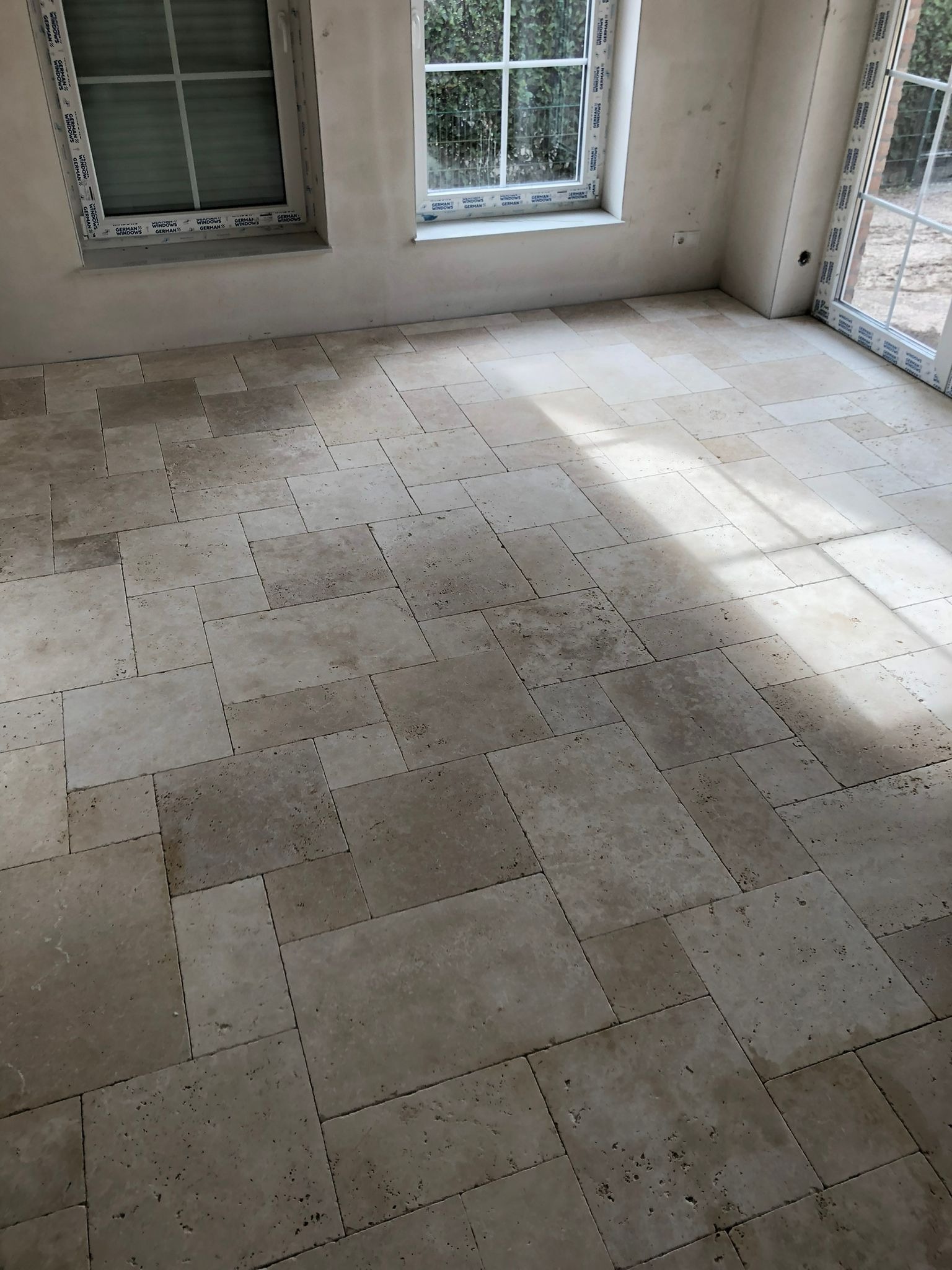 Ułożona podłoga z płytek trawertynowych w pomieszczeniu z dwoma oknami, ściany w stanie surowym, widoczny cień padający przez okno.