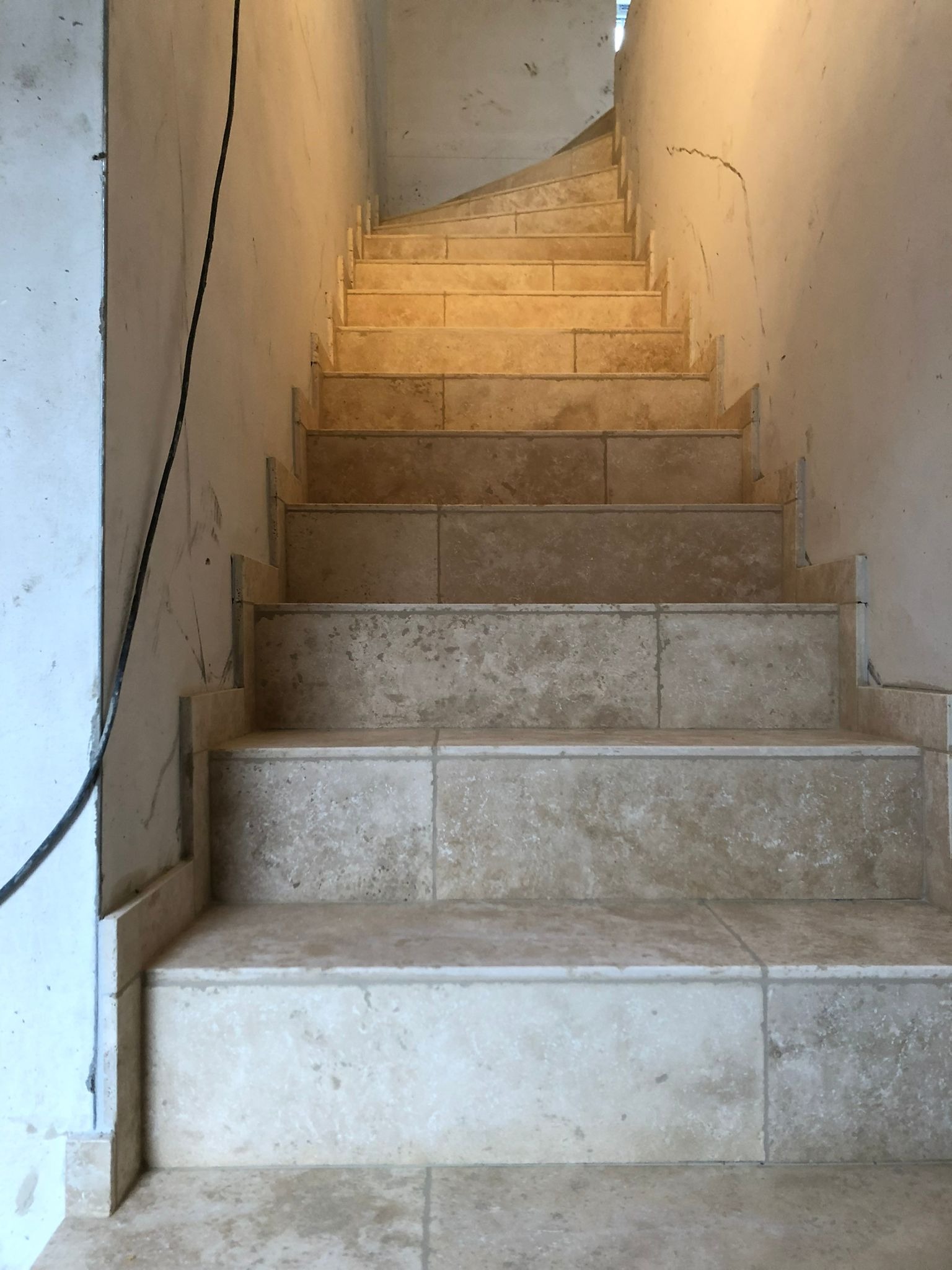 Schody wyłożone beżowymi płytkami imitującymi kamień, widoczne niedokończone ściany i wystający kabel elektryczny.