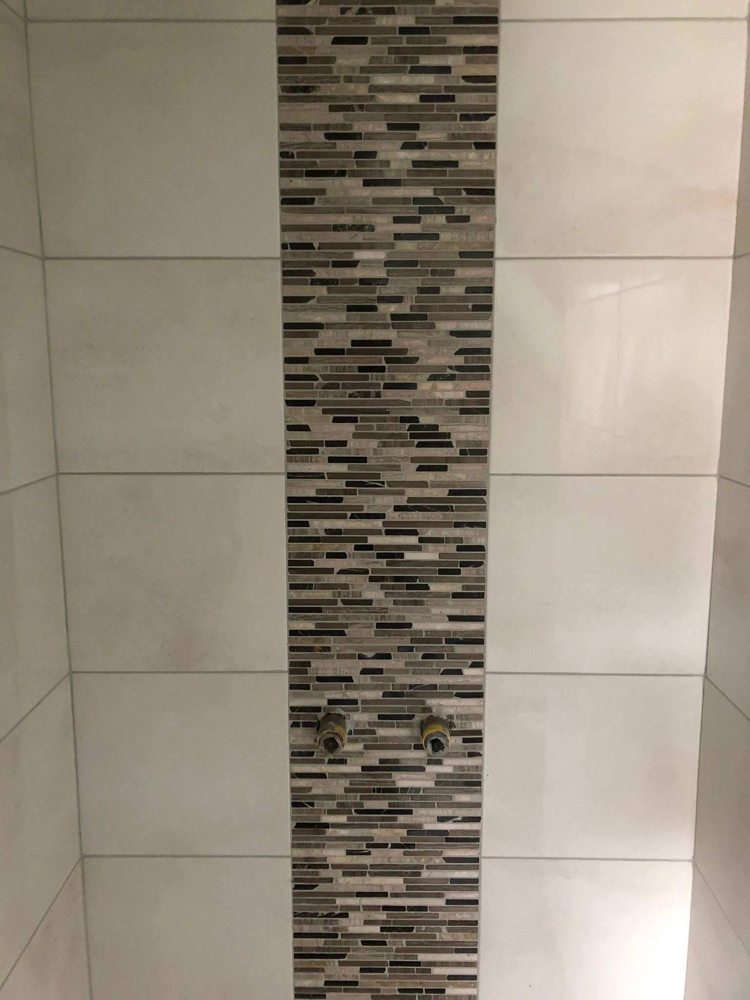 Fragment ściany w łazience z białymi, poziomymi płytkami i pionowym pasem mozaiki w odcieniach brązu i szarości, z widocznymi wystającymi rurami instalacji wodnej.