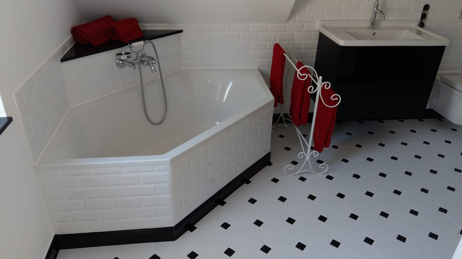 Biała, sześciokątna wanna obłożona płytkami typu metro, z chromowaną baterią i czerwonymi ręcznikami na półce oraz na ozdobnym wieszaku, na podłodze białe płytki z czarnymi rombami, czarna szafka...