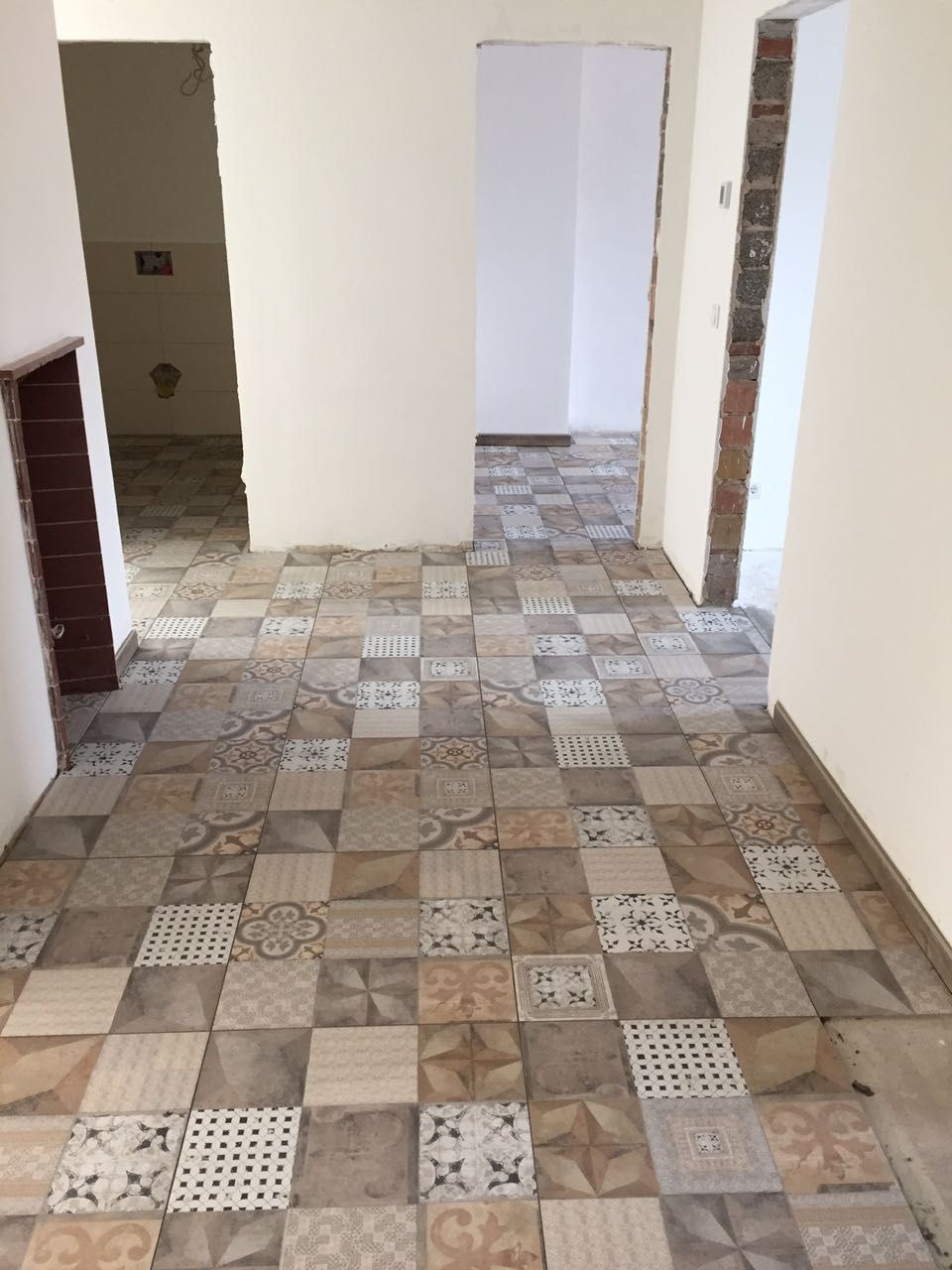 Ułożona podłoga z patchworkowych płytek ceramicznych w odcieniach beżu i brązu, widok na korytarz z otwartymi przejściami do innych pomieszczeń w trakcie remontu.