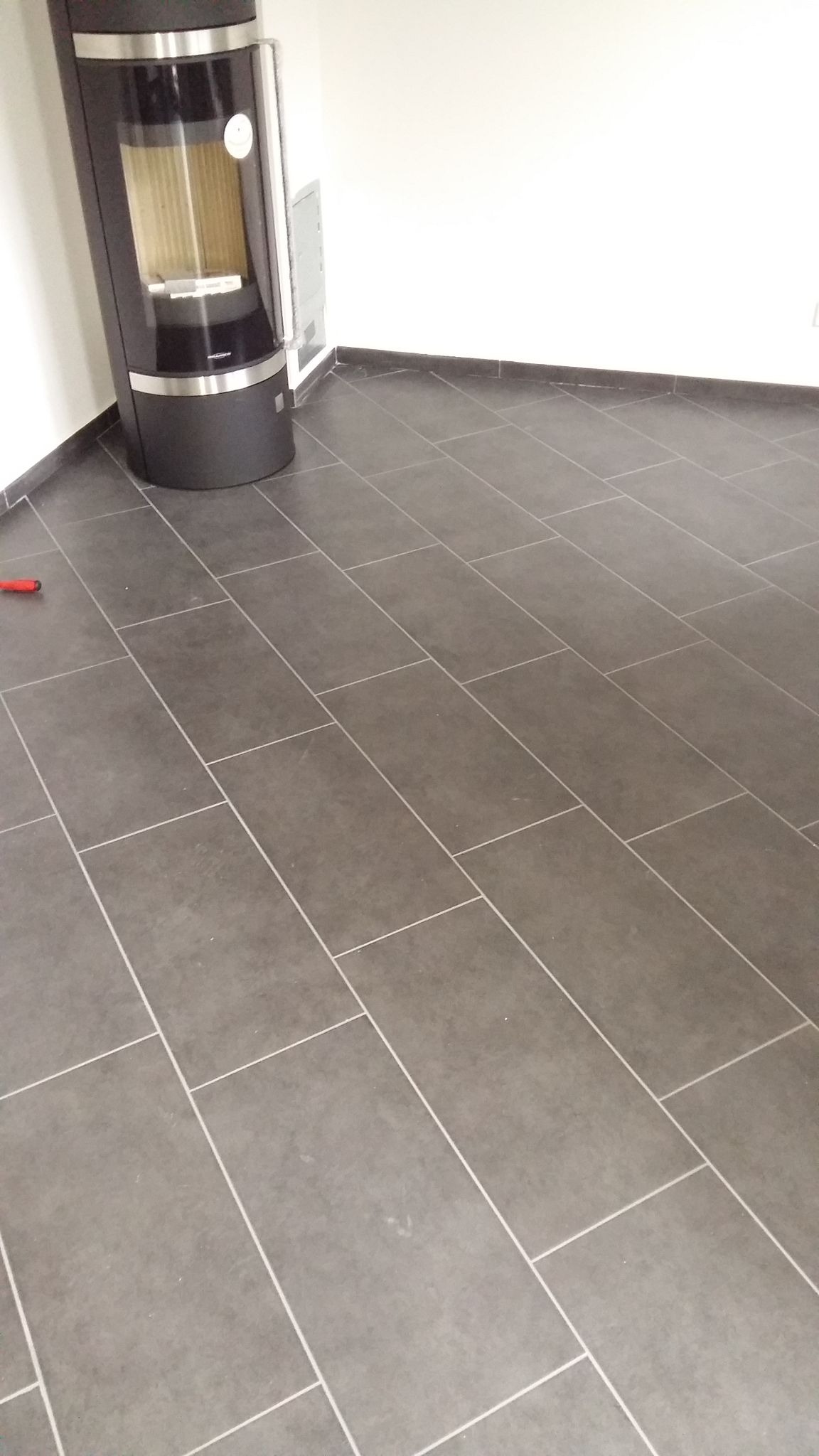 Ułożona podłoga z szarych, prostokątnych płytek gresowych z jasnoszarą fugą, obok stoi czarny piec kominkowy.