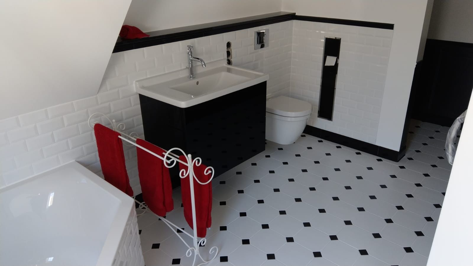 Wykończona łazienka w stylu retro z białą armaturą, czarnymi akcentami i podłogą w biało-czarną szachownicę. Na wieszaku dekoracyjne czerwone ręczniki.