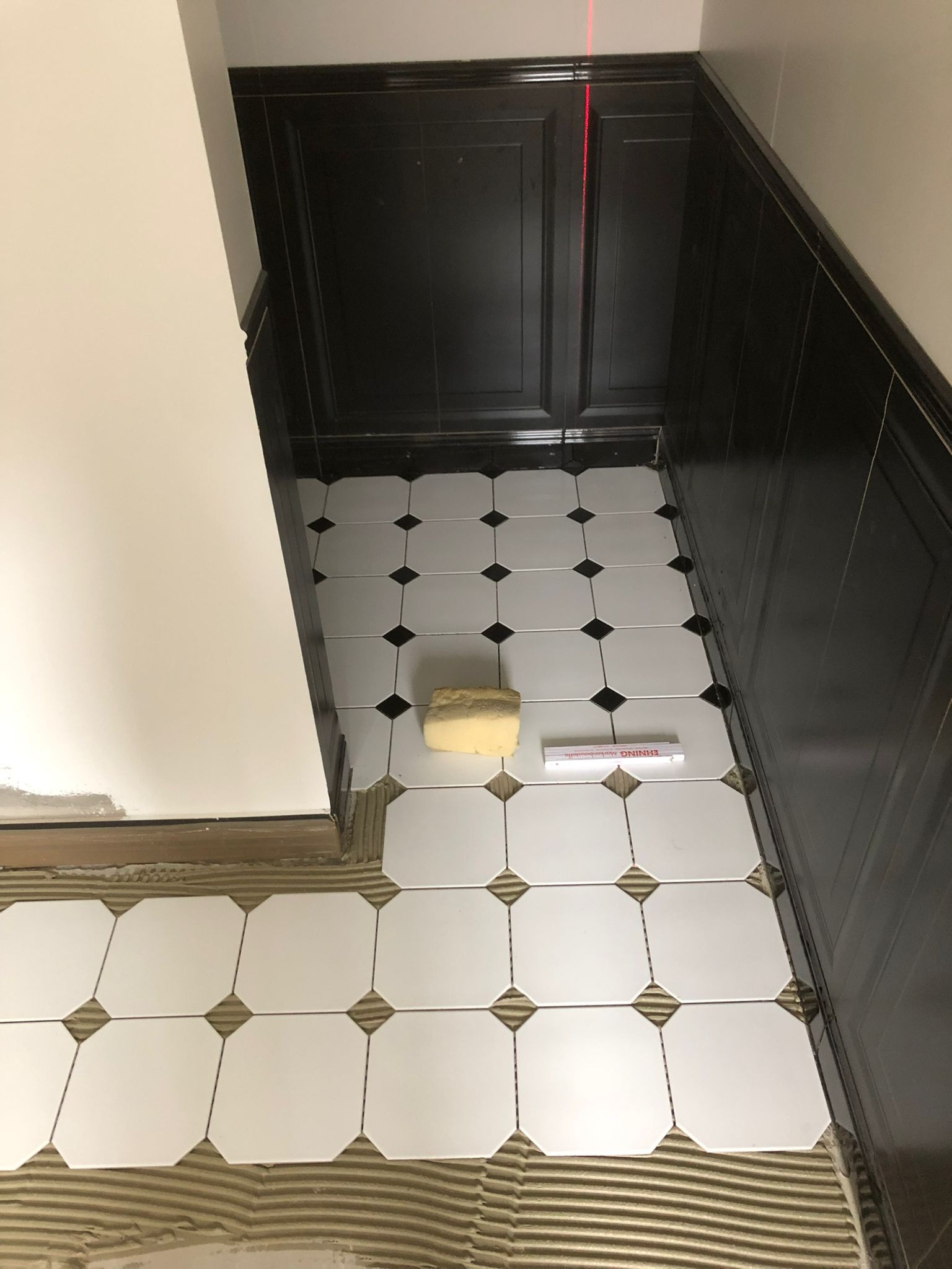 Układanie białych, ośmiokątnych płytek podłogowych z czarnymi wstawkami w narożach, widoczna poziomica i gąbka, w tle czarne płytki ścienne i czerwona wiązka lasera poziomującego.