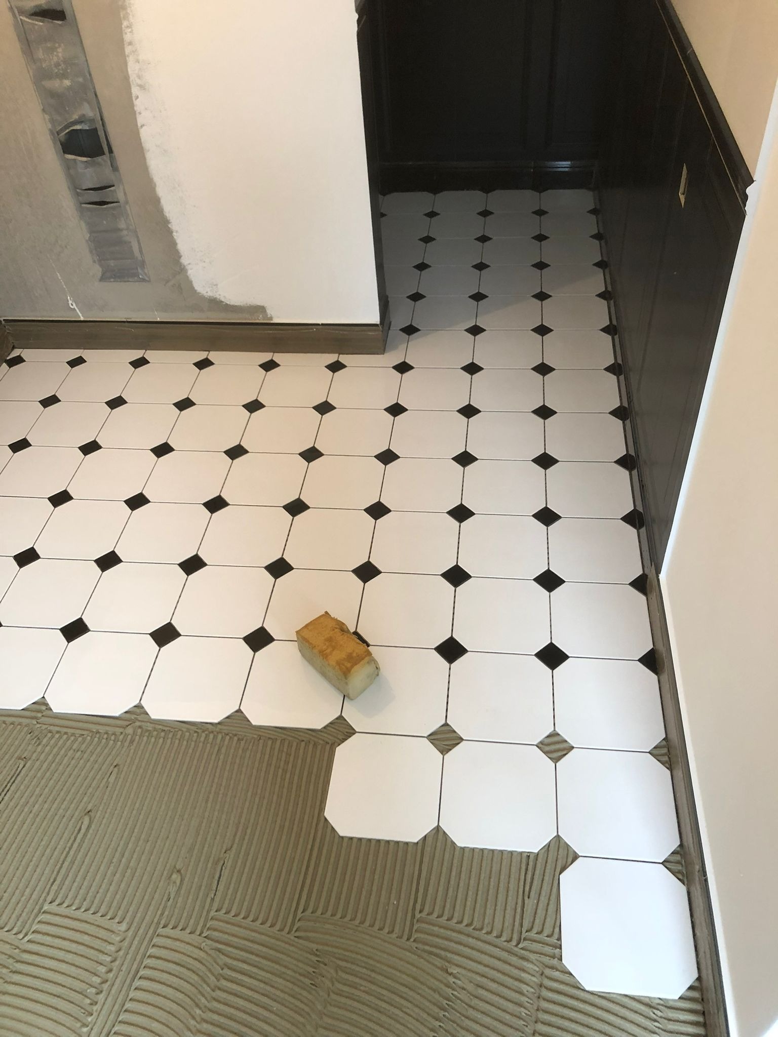 Układanie białych, ośmiokątnych płytek z czarnymi wstawkami na podłodze, widoczna warstwa kleju i gąbka do czyszczenia fug.