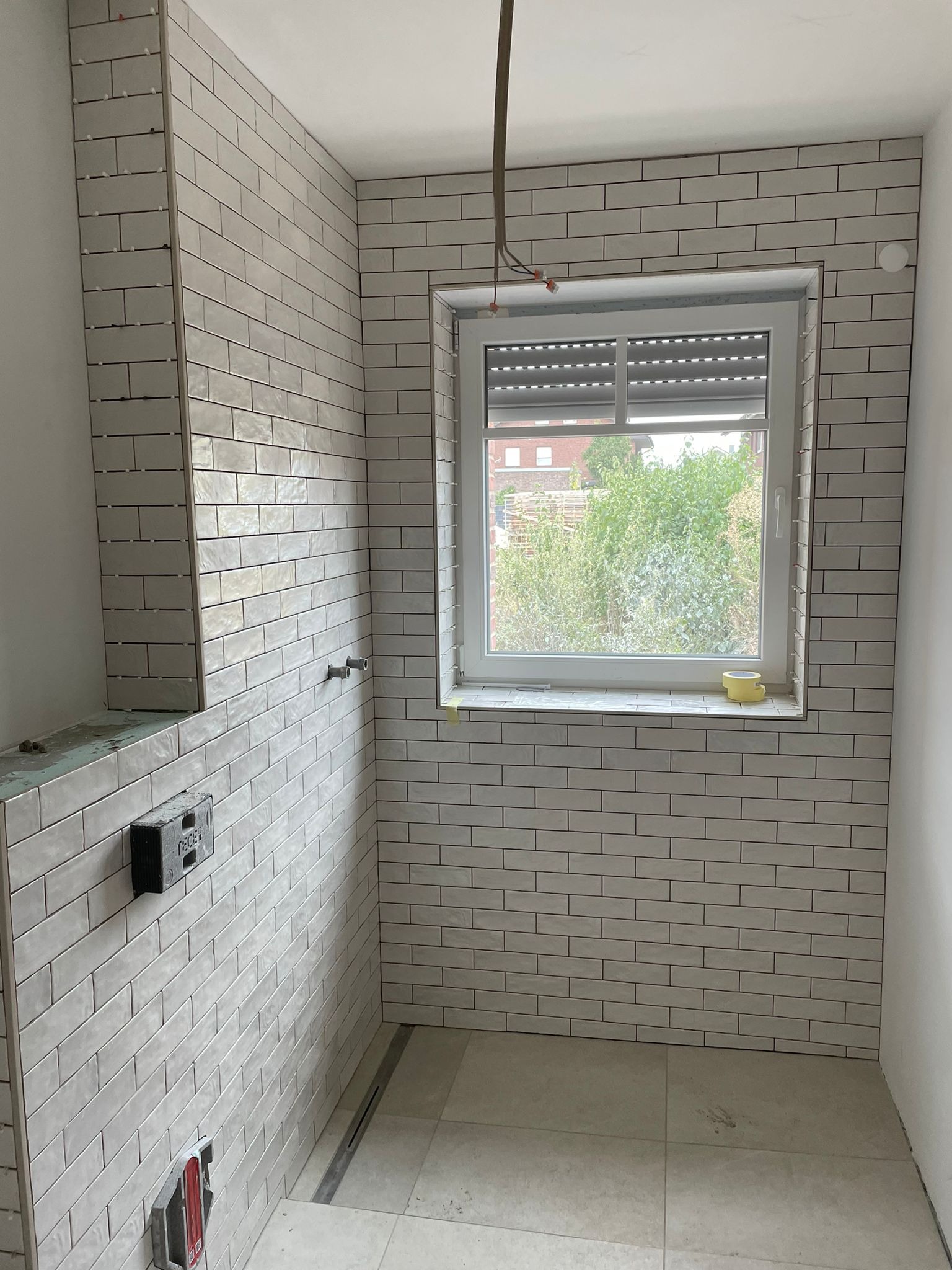 Wnętrze łazienki w trakcie układania białych, podłużnych płytek na ścianach, widoczne okno z roletą, fragment podłogi z jasnych płytek, oraz niedokończone elementy instalacji hydraulicznej.