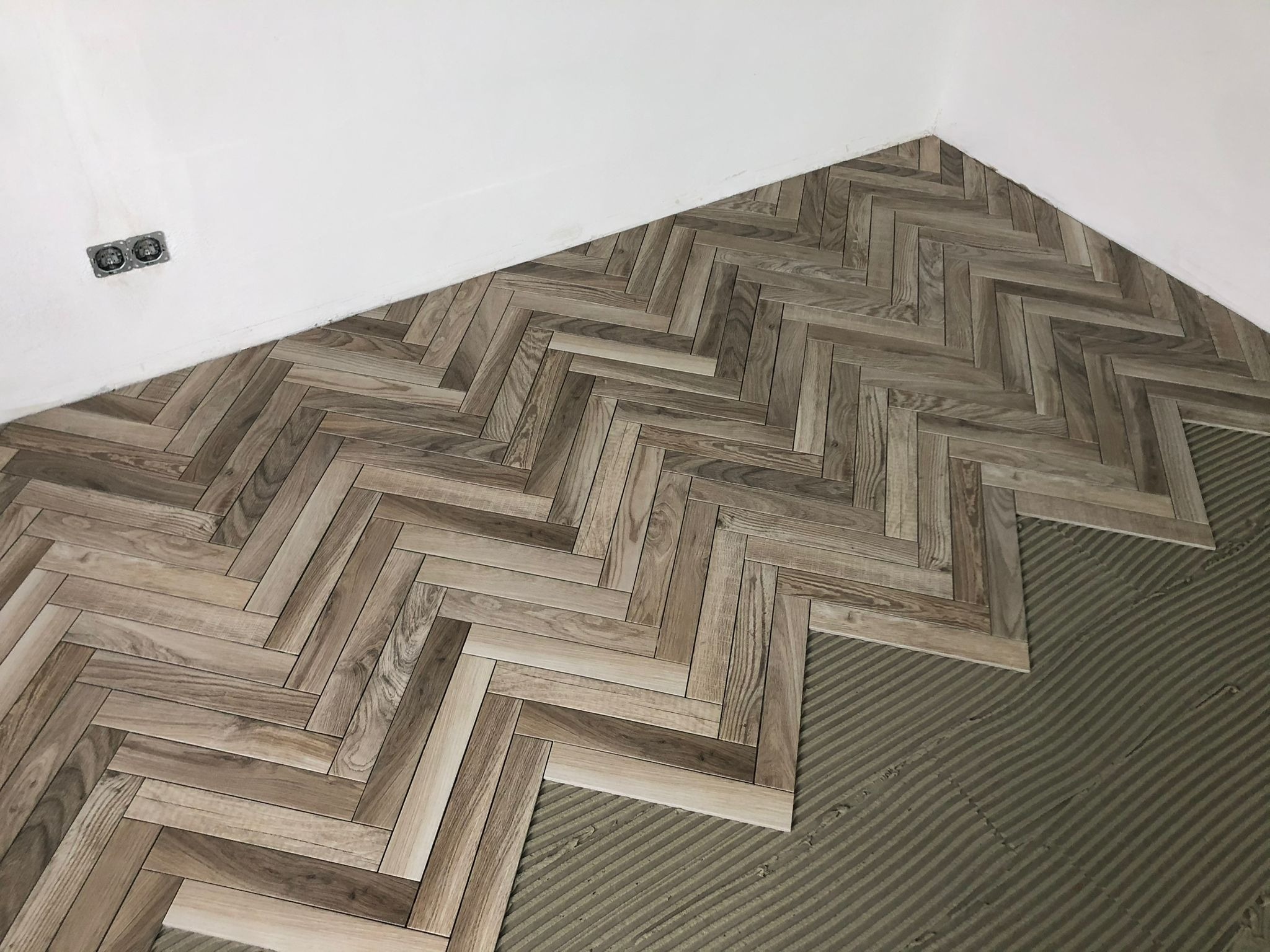Układanie parkietu ceramicznego w jodełkę, widoczny fragment podłogi z nałożonym klejem i ułożonym wzorem, białe ściany w tle, gniazdko elektryczne po lewej stronie.
