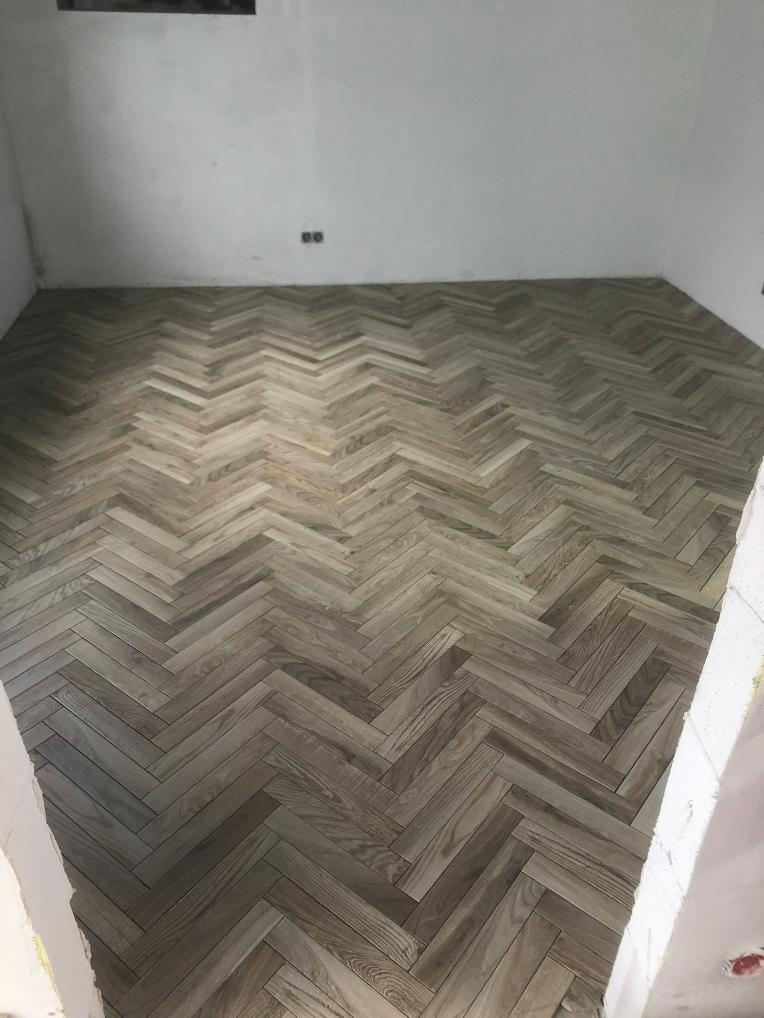 Ułożona podłoga z parkietu w jodełkę w nowo wyremontowanym pomieszczeniu, widoczne gniazdka elektryczne i surowe ściany.