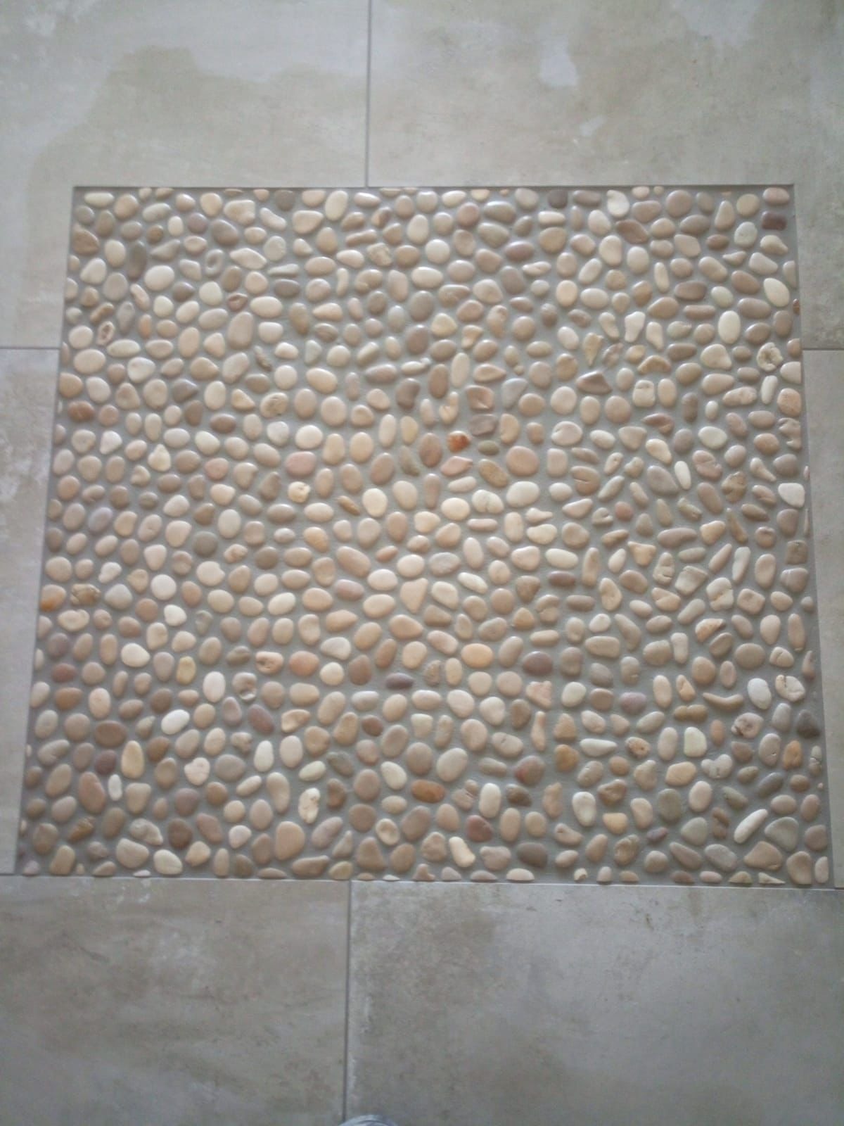 Fragment podłogi z dekoracyjnym wzorem z drobnych, okrągłych kamieni w odcieniach beżu i brązu, otoczonymi przez większe, kwadratowe płytki w szarym odcieniu.