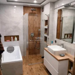 Nowoczesna łazienka z kabiną prysznicową typu walk-in, drewnopodobnymi płytkami na podłodze i ścianie prysznica, białą armaturą i szafką pod umywalką z blatem w kolorze drewna, oraz wnęką z półkami.