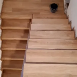 Drewniane schody w trakcie montażu, widoczne niedociągnięcia przy ścianie, podest wyłożony panelami, czarne wiadro z farbą w rogu podestu.