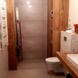 Wnętrze łazienki w trakcie remontu, widoczna szara płytka na ścianach i podłodze, drewniane obudowy pionów, biała toaleta, poziomica oparta o ścianę, butelka wody na blacie umywalki.