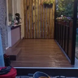 Zadaszony taras z drewnianą ścianą i terakotą na podłodze, oświetlony od góry, z widokiem na ogród w Zamościu. Na pierwszym planie wiadro i pojemnik z farbą.