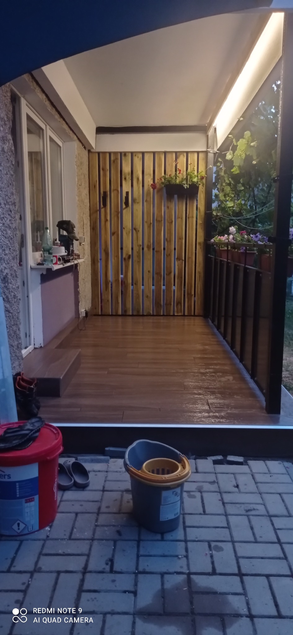 Zadaszony taras z drewnianą ścianą i terakotą na podłodze, oświetlony od góry, z widokiem na ogród w Zamościu. Na pierwszym planie wiadro i pojemnik z farbą.