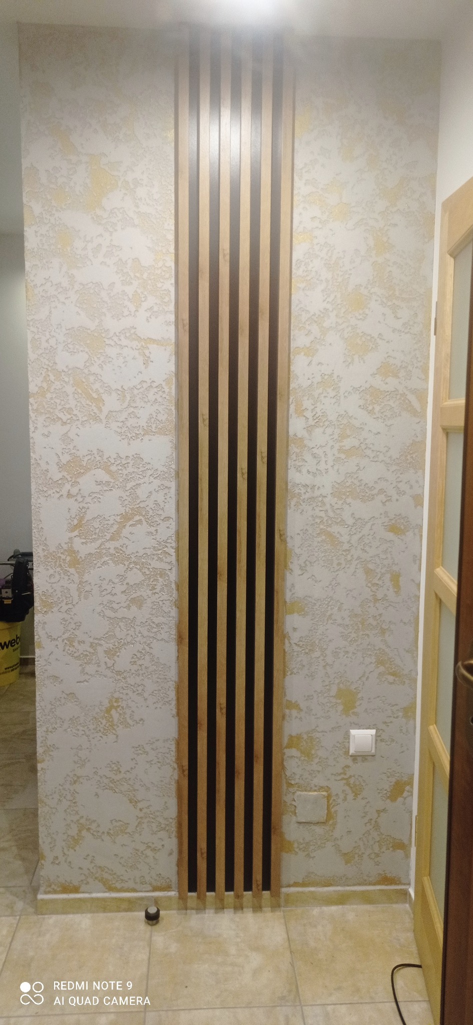 Ściana z dekoracyjnym tynkiem w odcieniach bieli i złota, z pionowymi panelami drewnianymi w kolorze jasnego i ciemnego brązu, tworzącymi nowoczesny akcent.