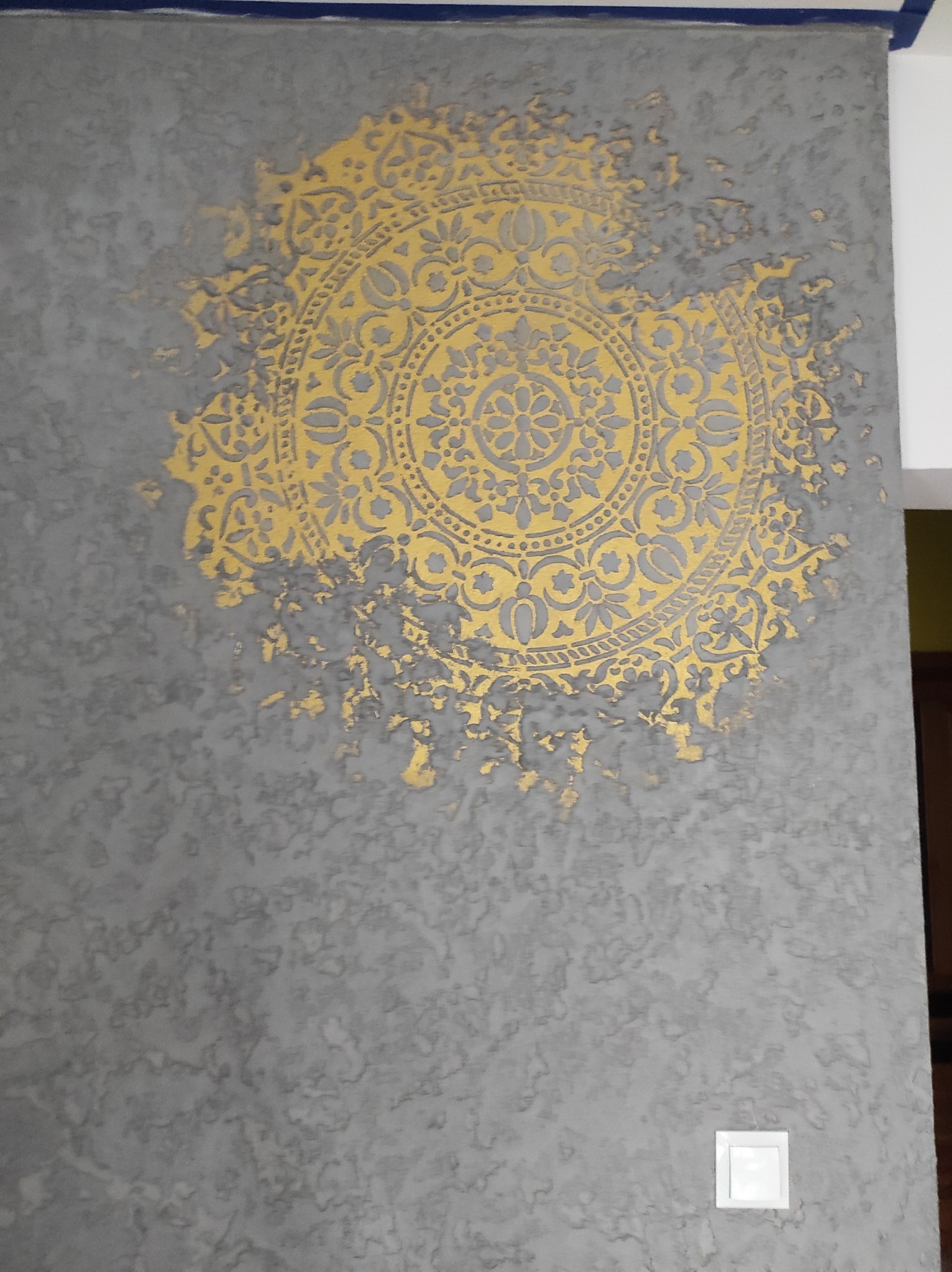 Szara ściana z dekoracyjnym, złocistym wzorem mandali, częściowo z efektem spękania i postarzenia.