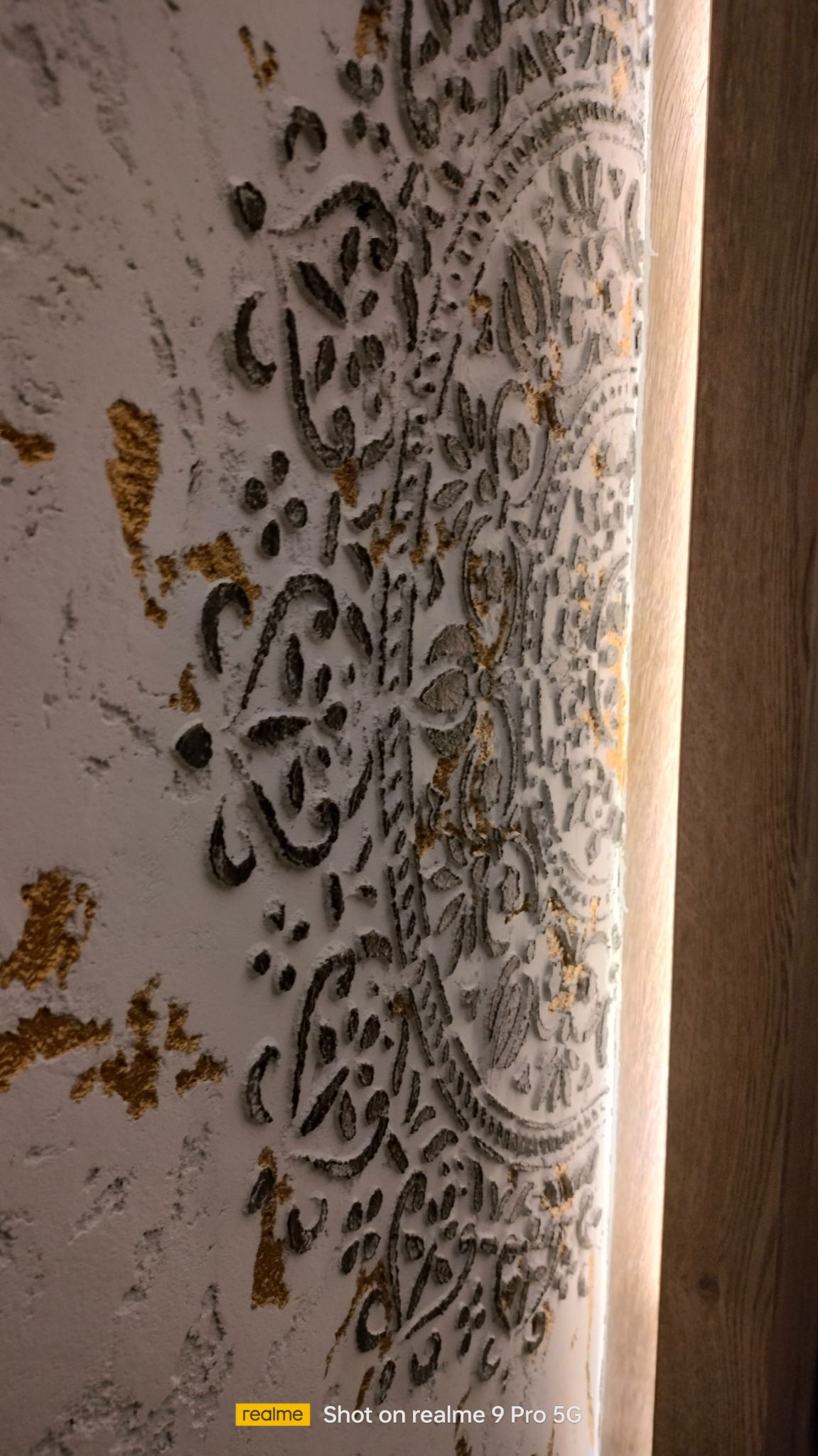 Dekoracyjna ściana z trójwymiarowym wzorem ornamentu w odcieniach bieli, szarości i złota, wykonana techniką stiuku weneckiego, widoczna z boku, z fragmentem drewnianej boazerii w tle.