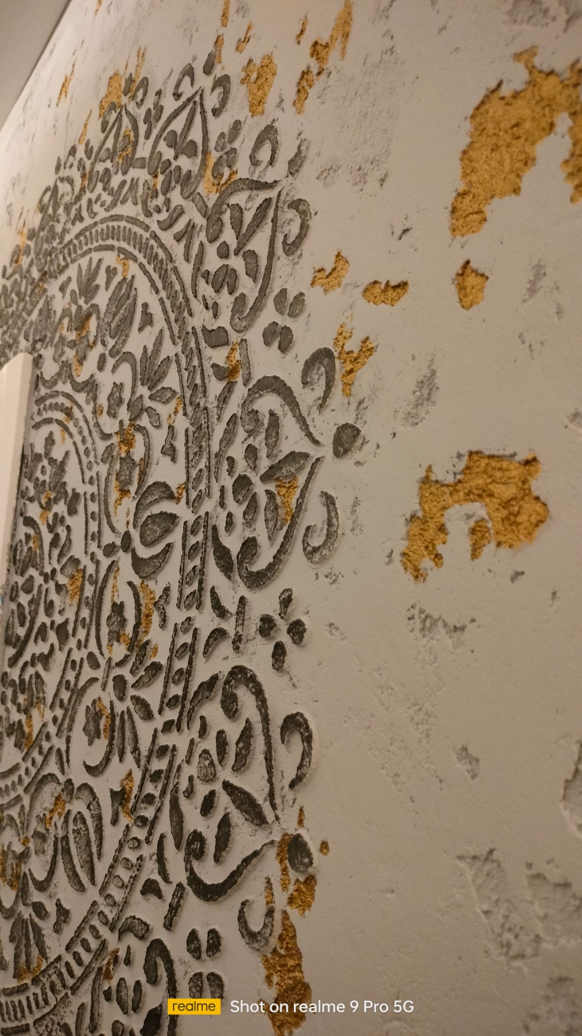 Dekoracyjna ściana z szarym wzorem mandali i złotymi akcentami, nierówna faktura tynku.