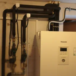 Biała pompa ciepła Panasonic Aquarea Smart Cloud z czarnymi rurami instalacji grzewczej i rozdzielnicą elektryczną na ścianie w pomieszczeniu technicznym.