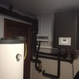 Szare urządzenie grzewcze marki Stiebel Eltron zamontowane na ścianie z beżowych płytek, obok biały zbiornik z manometrem i czarne rury instalacyjne.