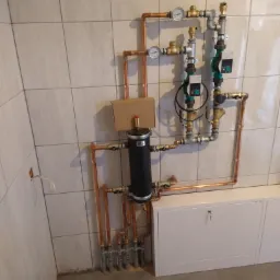 Instalacja hydrauliczna z miedzianych rur, czarny zbiornik z kartonowym pudełkiem na górze, manometry i pompy obiegowe zamontowane na ścianie z jasnych płytek.