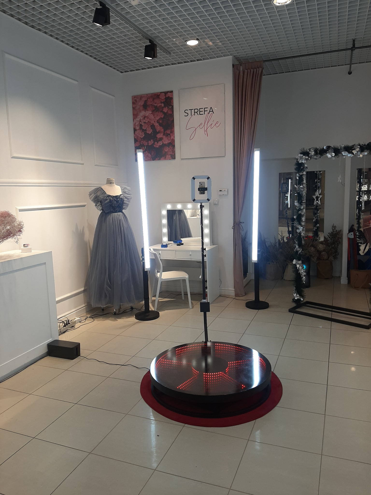 Oświetlona platforma obrotowa z telefonem, otoczona lampami LED, w tle manekin w długiej sukni, toaletka z lustrem i dekoracje.