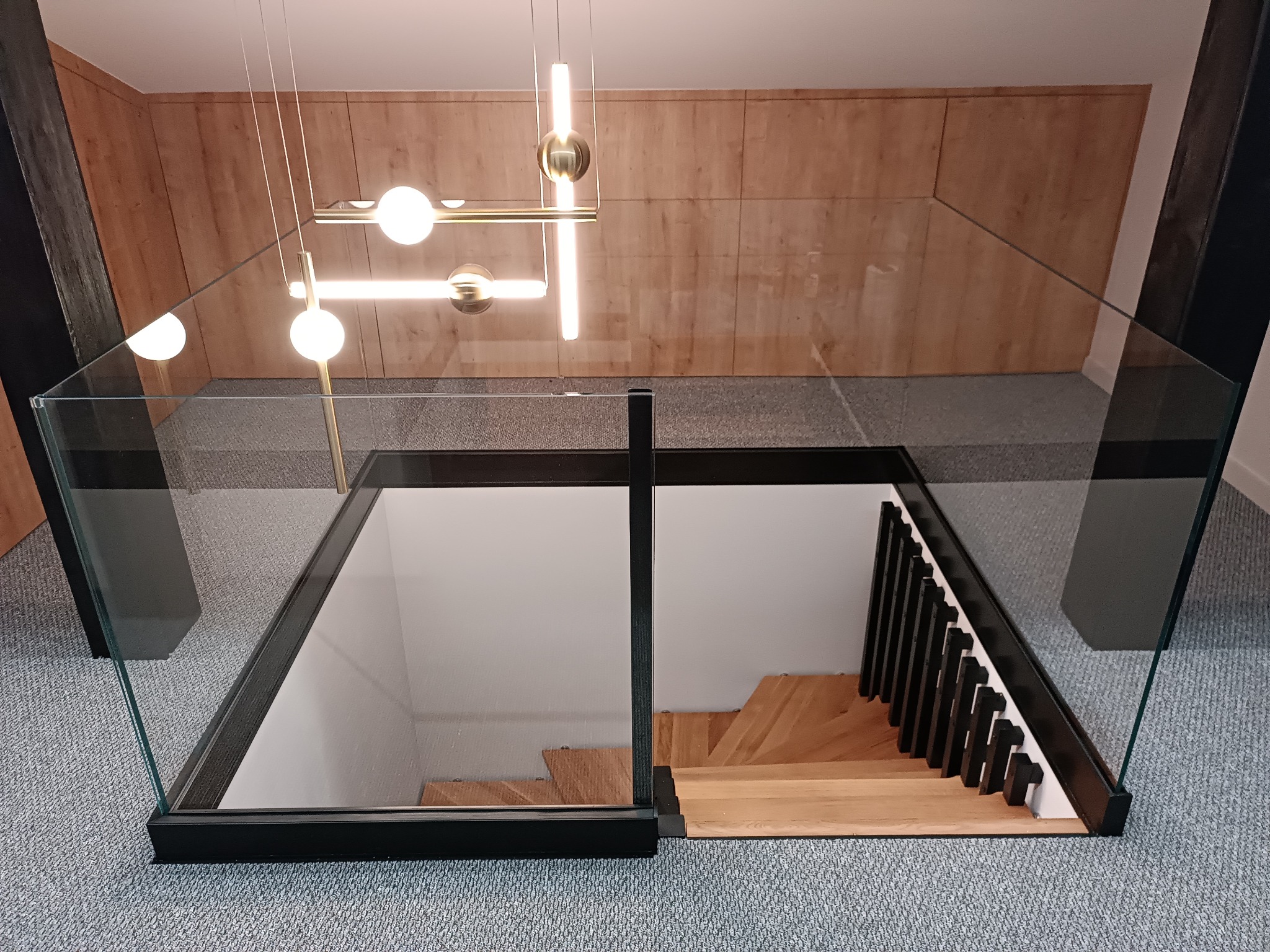Szklana bariera zabezpieczająca schody z drewnianymi stopniami i czarną poręczą, widok z góry, nowoczesne oświetlenie wiszące.
