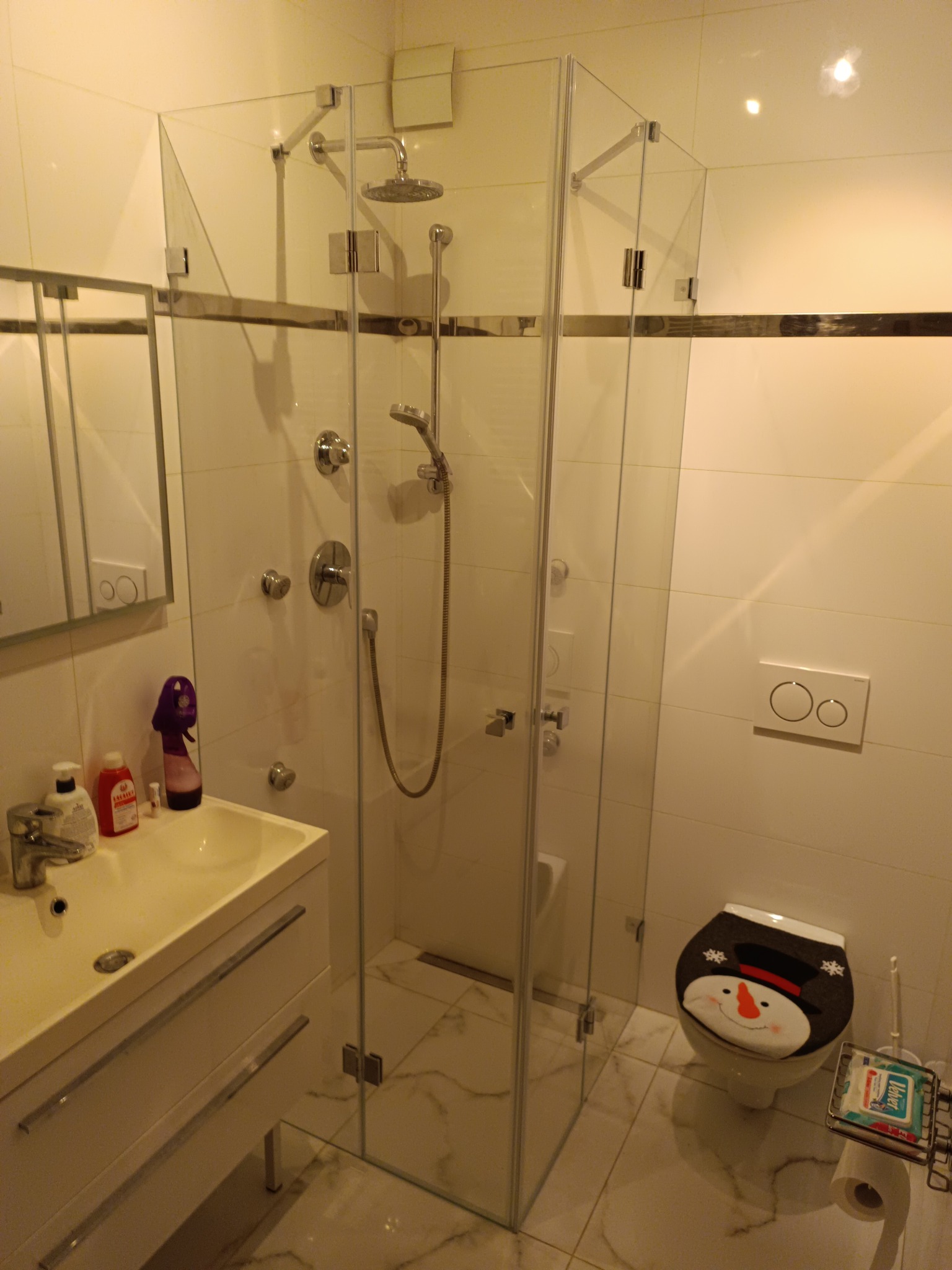 Szklana kabina prysznicowa z chromowanymi okuciami w jasnej łazience z marmurową podłogą i toaletą z nakładką w kształcie bałwana.