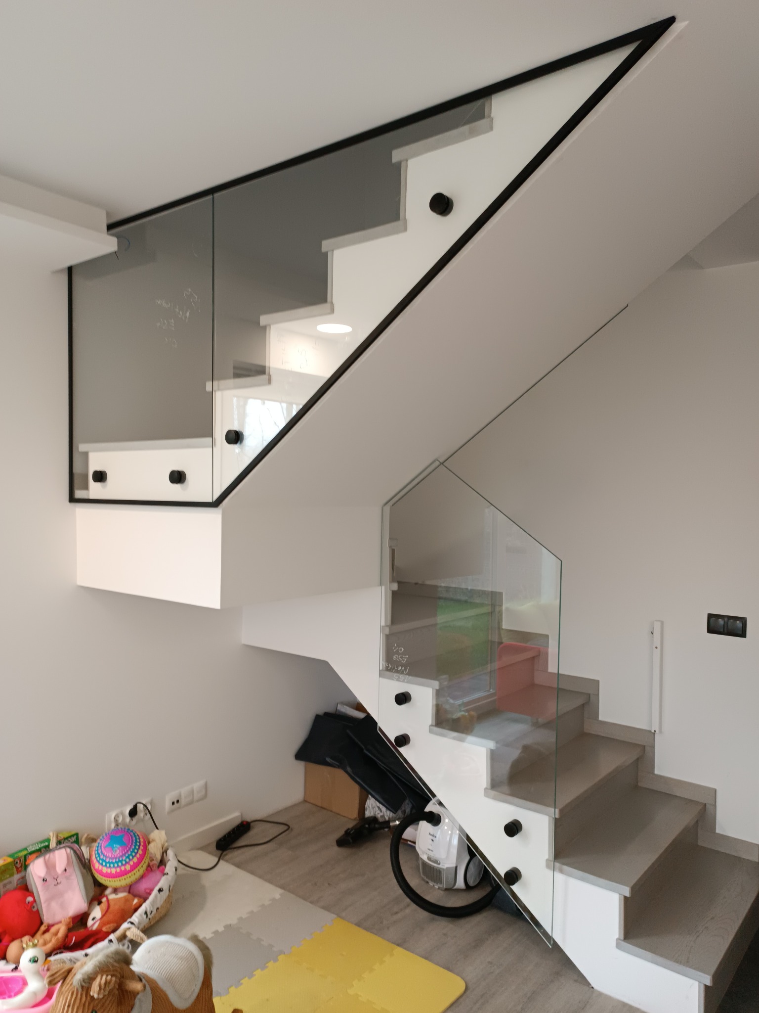 Nowoczesne schody z balustradą szklaną mocowaną punktowo, czarne detale wykończeniowe, widok z dołu na jasne stopnie i fragment pokoju z zabawkami.