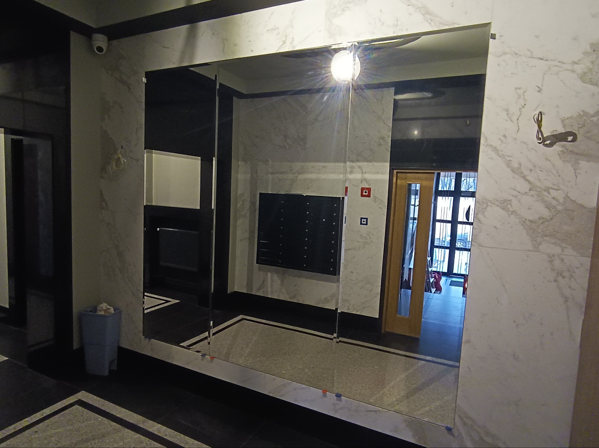 Duże lustro na ścianie w holu wejściowym budynku, odbijające skrzynki pocztowe, drzwi wejściowe i fragment grzejnika.