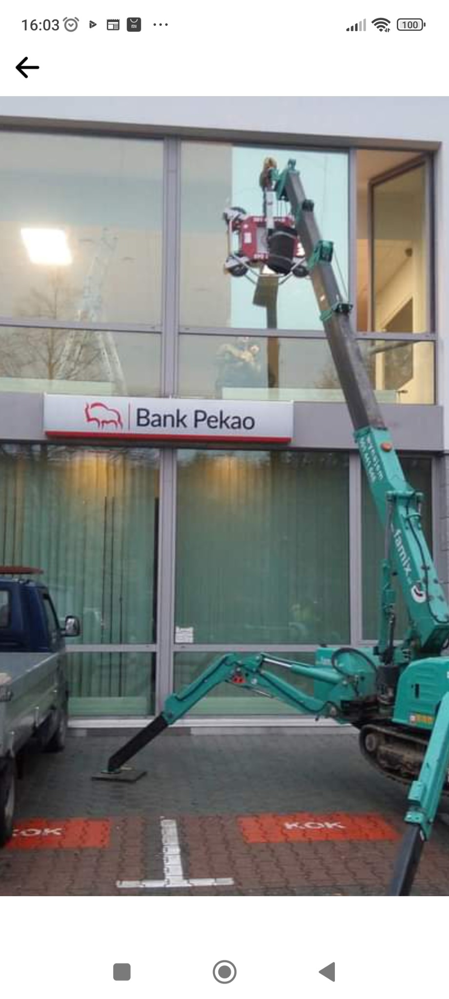 Montaż okien w budynku banku z użyciem podnośnika teleskopowego z przyssawkami; widoczny fragment elewacji, okna i logo Banku Pekao.