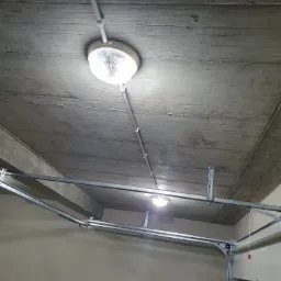 Dwie lampy sufitowe z białymi kloszami, zamontowane na szarym, betonowym suficie garażu, widoczne szyny prowadzące bramę garażową.