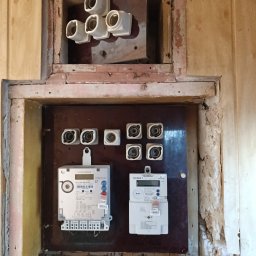 VIBEELEKTRYK ADRIAN KLISZ - Skrzynka z dwoma licznikami elektrycznymi na brązowej płycie, powyżej bezpieczniki topikowe, ściany obudowy w trakcie remontu, widoczne narzędzia elektryczne.
