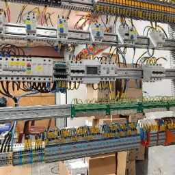 Rozdzielnia elektryczna z licznymi bezpiecznikami, przewodami oznaczonymi żółtymi etykietami i listwami zaciskowymi, zamontowana na metalowej konstrukcji w pomieszczeniu technicznym.