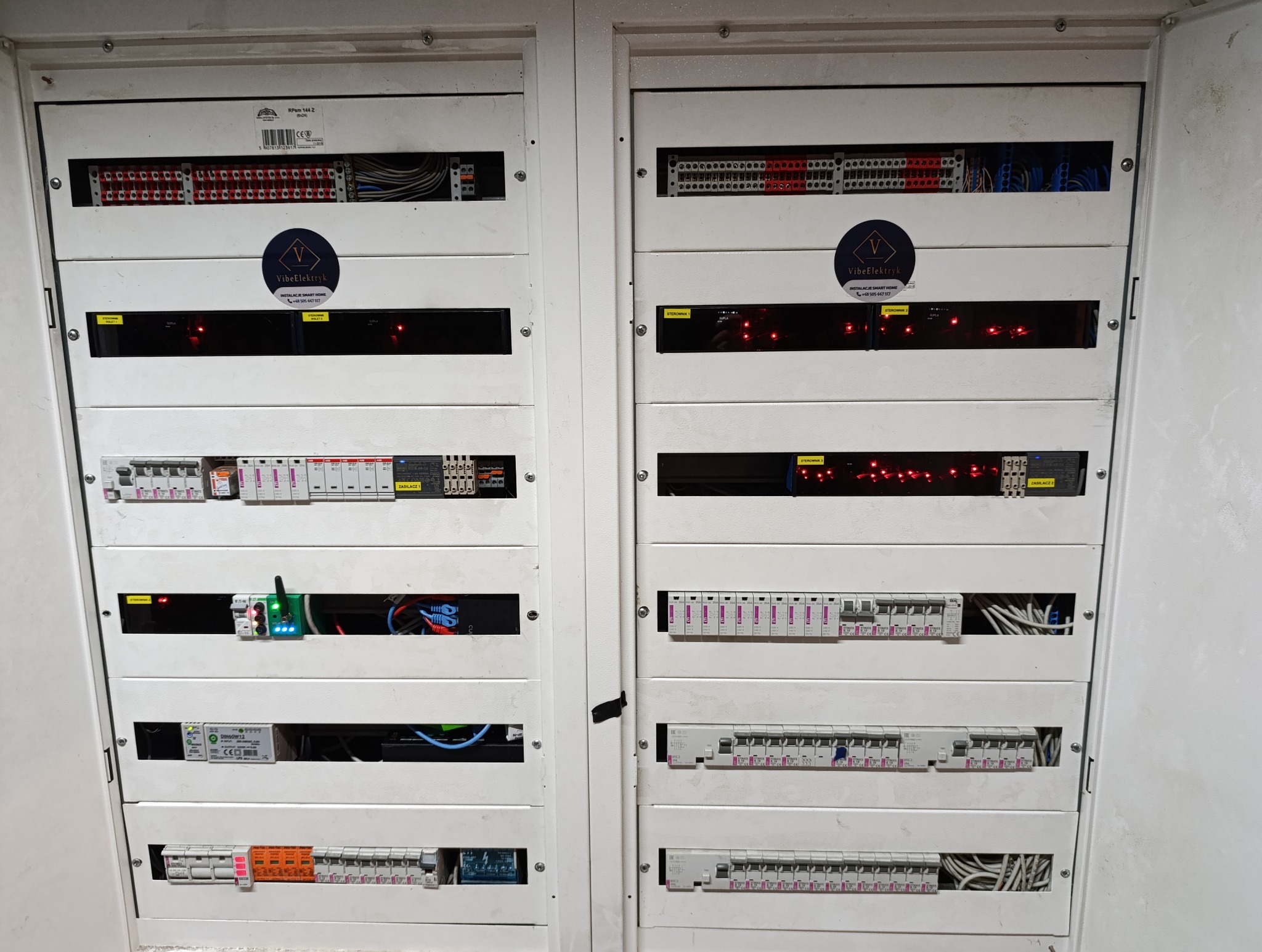 Dwa panele elektryczne z różnymi zabezpieczeniami, sterownikami i okablowaniem, widoczne naklejki z logo firmy VibeElektryk oferującej instalacje smart home.